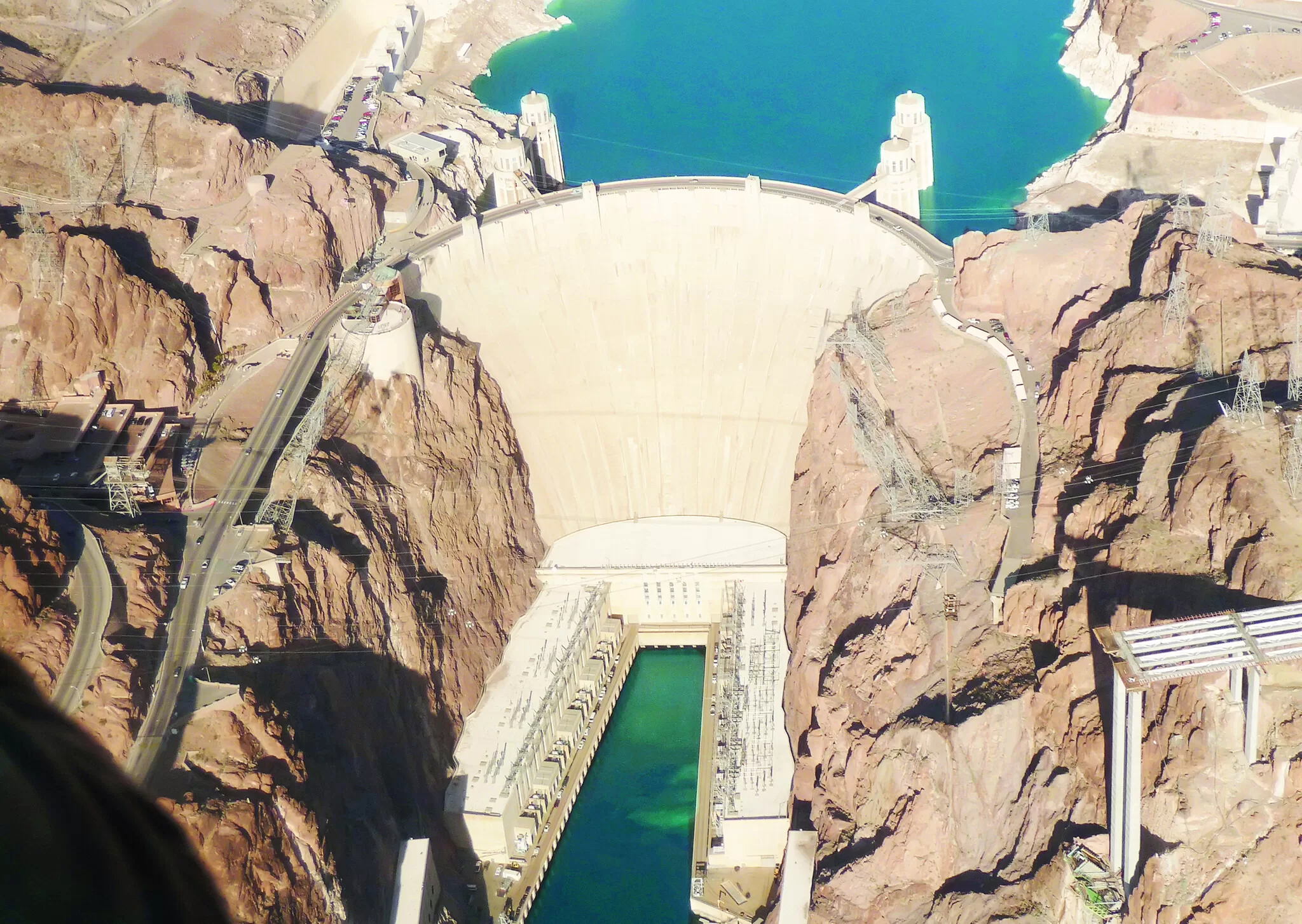 Luftaufnahme des Hoover-Damms inmitten roter Felsen, mit türkisfarbenem Wasserreservoir und Infrastruktur rund um den Damm.
