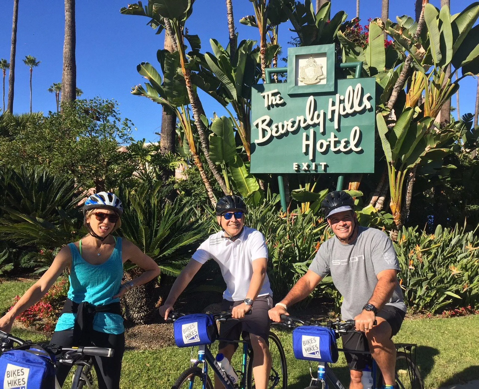 Mensen staan met fietsen voor het bord van het Beverly Hills Hotel, omringd door groene natuur op de achtergrond