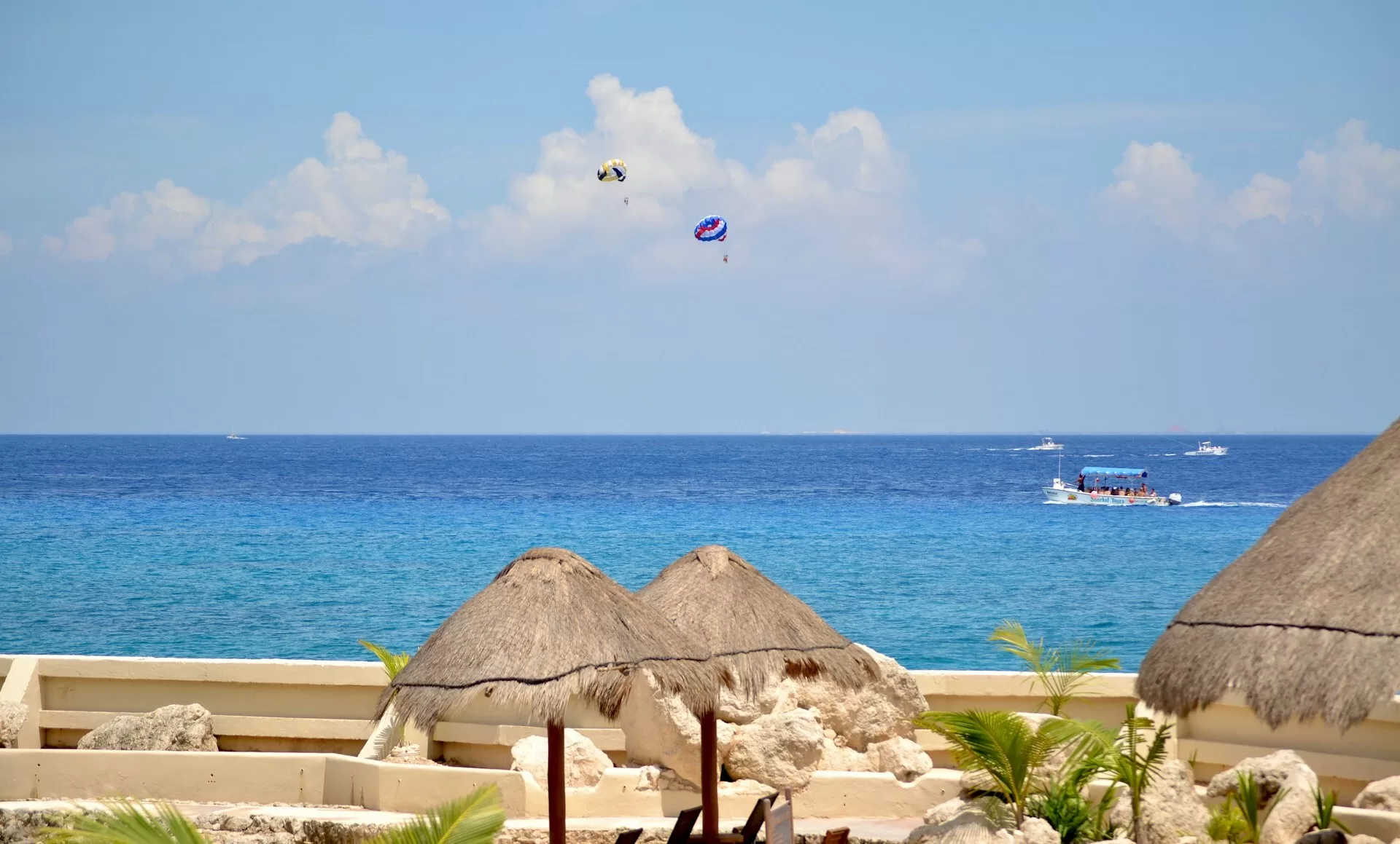 Rieten parasols op een aangelegd terras voor de kust van Cozumel met parasailing op de achtergrond.